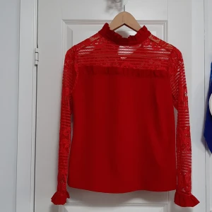 Röd blus med spets och volanger - Säljer en röd blus med högt krage och volangdetaljer. Blusen har långa ärmar i transparent spets med blommigt mönster och volang vid ärmslut. Ovanför bröstet finns en dekorativ volang och spetsen fortsätter över axlarna. Perfekt för dig som gillar statement-plagg.