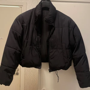 Svart croppad pufferjacka - Säljer en svart croppad pufferjacka med hög krage och dragkedja framtill. Jackan har långa ärmar, två sidofickor och snörning i nederkant för att justera passformen. 