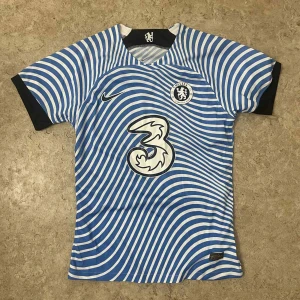 Chelsea FC blå randig fotbollströja Nike - Chelsea FC fotbollströja från Nike med ett unikt blått och vitt vågigt randigt mönster. Kortärmad modell med svarta detaljer vid ärmslut och hals. Tryckt klubbmärke och sponsorlogga på bröstet. Tillverkad i lätt och ventilerande polyester.