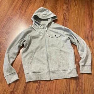 Grå hoodie från Polo Ralph Lauren - Snygg grå hoodie från Polo Ralph Lauren med dragkedja och huva med vita snören. Tröjan har två fickor framtill och en liten svart Polo-logga broderad på bröstet. Perfekt för chill dagar och enkel att matcha med jeans eller joggers.