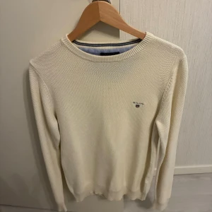 Vit stickad tröja från GANT - Vit stickad tröja från GANT med rund hals och diskret logga broderad på bröstet. Tröjan har långärmad passform och ribbade muddar vid ärmslut och nederkant. Perfekt för dig som gillar klassisk och clean stil.