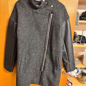 Gråsvart kappa från Noisy May, stl L - Snygg gråsvart kappa från Noisy May i storlek L. Jackan har coola svarta skinnärmar, sned dragkedja och hög krage med tryckknappar. Materialmix av ull och skinn ger en edgy vibe. Perfekt för dig som vill sticka ut med en modern look.
