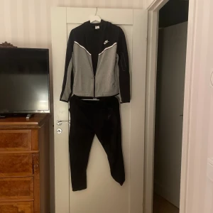 Nike hoodie svart och grå XL - Nike hoodie och byxor i svart och grått med vit detalj över bröstet. Tröjan har dragkedja, huva och långa ärmar. Perfekt för träning eller chill. Klassisk Nike-logga på bröstet och bekväm passform. Dock ett litet hål drygt 1cm i vänstra ficka men de syns ej när man använder den.