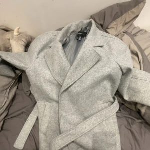 Ljusgrå kappa från Vero Moda XS - Snygg ljusgrå kappa från Vero Moda i storlek XS. Klassisk modell med bred krage och bälte i midjan för en clean och stilren look. Perfekt för dig som gillar minimalistisk stil och vill ha en jacka som funkar till många outfits.