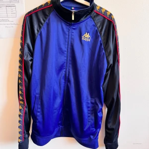 Blå och svart track jacket från Kappa - Snygg blå och svart track jacket från Kappa med gul logga på bröstet och klassiska Kappa-band längs ärmarna. Jackan har hel dragkedja, ståkrage och är tillverkad i 100% polyester. Röd detalj längs axlarna och fickor på sidorna.