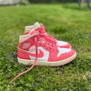 Nike Air Jordan 1 rosa/vita sneakers - Nike Air Jordan 1 sneakers i en snygg kombo av rosa och vitt med klassisk hög siluett. Skorna har rosa snörning, vit tåbox och rosa swoosh. Ovandelen är i skinn och textil, med perforerad tå för extra ventilation. Perfekta för dig som vill sticka ut med färg.