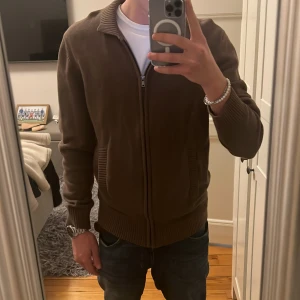 Brun kofta med dragkedja från Dressmann - Säljer en brun kofta från Dressmann i storlek M. Koftan är stickad med ribbade muddar, krage och fickor samt hel dragkedja framtill. Perfekt för lager-på-lager och passar till jeans eller chinos.