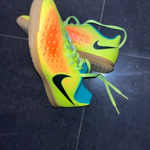 Nike fotbollsskor i neon och orange - Säljer ett par grymma Nike fotbollsskor i neon-gult med orange detaljer och svart swoosh. Skorna har snörning, platt gummisula och turkos insida. Perfekta för inomhusspel med cool färgkombination och bekväm passform.