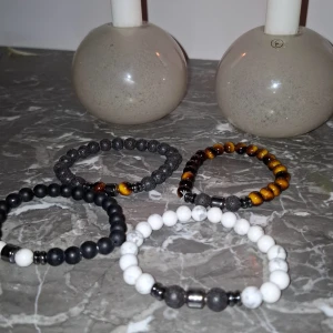 Fyra pärlarmband i olika färger - Säljer ett set med fyra snygga pärlarmband. Ett är helsvart med matta pärlor, ett är vitt med grå marmorering, ett är mörkgrått med två bruna pärlor och ett är brunt med tigeröga-effekt. Alla har rund form och elastisk tråd. Perfekt för att mixa och matcha till din stil.
