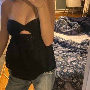 Svart axelbandslös tubtopp med broderi  - Säljer en svart axelbandslös tubtopp med snygg broderad spets och ett litet cutout-detalj framtill. Toppen har bandeau-formad överdel och är luftig nedtill, perfekt till jeans eller kjol. Den är ärmlös och har en cool, ungdomlig vibe.