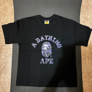 Svart BAPE t-shirt med camo-print - Svart t-shirt från BAPE med klassiskt camo-print i blå och lila toner på bröstet och stor logga 'A Bathing Ape'. Rund hals och korta ärmar. Ikonisk BAPE-tag på ärmen. Tillverkad i mjuk bomull, perfekt för streetwear.