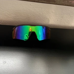 Sportiga regnbågssolglasögon från Velo - Säljer ett par sportiga solglasögon från Velo med spegelglas i regnbågsfärger. Modellen har en bred, böjd skärm och svarta skalmar i syntetmaterial. Perfekt för dig som vill ha en cool look när du tränar eller är ute i solen.
