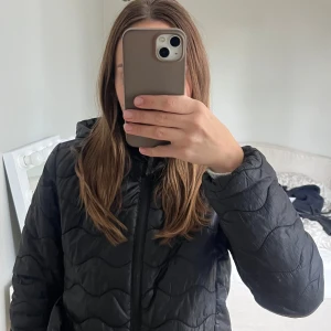 Svart pufferjacka från Samsa Collection - Svart pufferjacka från Samsa Collection med vågigt quiltat mönster och huva. Jackan har dragkedja framtill och är i en klassisk, enkel modell som passar till många olika outfits. Perfekt för kyligare dagar. Den har två defekter (se bild 4 och 5 ) men funkar om den ska ❤️🤗passar även storlek xxs/xs