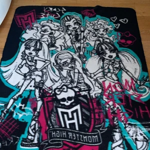 Monster High filt - Mjuk och varm fleecefilt med motiv av de klassiska Monster High-karaktärerna.Perfekt till soffan, sängen eller som samlarobjekt för fans av den ikoniska 2010-talseran. Snyggt tryck i svart, turkos, rosa och vitt – riktigt nostalgisk vibe!