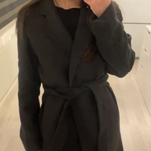 Svart kappa med bälte - Stilren svart kappa med bälte i midjan och klassisk krage. Långärmad modell med rak passform, perfekt för kyligare dagar. Enkel och tidlös design som passar till det mesta.