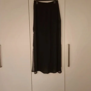 Svart långkjol från H&M DIVIDED - Svart långkjol från H&M DIVIDED i storlek S. Kjolen har ett tunt, transparent yttertyg med ett kortare innertyg som ger en snygg layering-effekt. Resår i midjan för skön passform. Perfekt för dig som gillar stilrena och enkla plagg med lite edge.