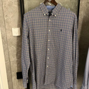 Rutig skjorta från Polo Ralph Lauren - Säljer en klassisk rutig skjorta från Polo Ralph Lauren i blått, beige och vitt. Skjortan har lång ärm, knappar hela vägen och button-down krage. Materialet är mjuk bomull och loggan är broderad på bröstet. Riktigt fet som overshirt och inga defekter!