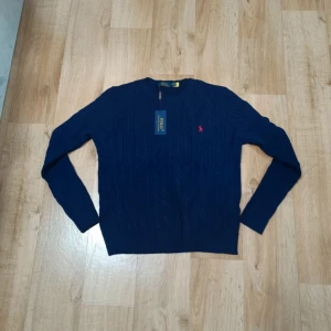 Marinblå stickad tröja Polo Ralph Lauren - Snygg marinblå stickad tröja från Polo Ralph Lauren med klassiskt kabelstickat mönster och röd broderad logga på bröstet. Tröjan har rund halsringning och långa ärmar, perfekt för en stilren look.