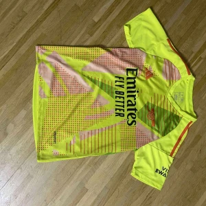 Arsenal x Adidas gul matchtröja - Arsenal fotbollströja från Adidas i neon-gult med röda och gröna detaljer. Tröjan har korta ärmar, tryckt klubbmärke och sponsorloggor samt ett unikt grafiskt mönster framtill. Tillverkad i lätt och ventilerande polyester, perfekt för träning eller match.