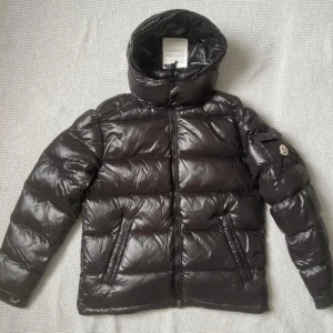 Moncler Maya - Helt ny, Snygg svart pufferjacka från Moncler med glansig finish och klassisk logga på ärmen. Jackan har huva, dragkedja framtill och två snedställda fickor. Insidan har ett coolt serietryck och märkestaggar. Perfekt för kalla dagar när du vill hålla stilen.