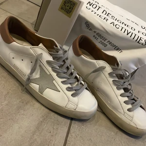 Golden Goose vita sneakers med grå stjärna - Golden Goose sneakers i vitt skinn med grå mockadetaljer och ikonisk stjärna på sidan. Skorna har grå snörning, bruna hälkappor i skinn och platt sula. Perfekta för dig som gillar en clean men ändå unik look.  Skorna är storlek 41 men jag skulle vilja säga att de passar 42 mycket bättre 
