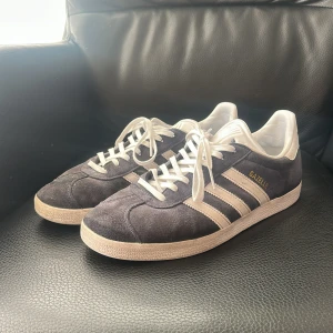 Adidas Gazelle mörkgrå sneakers - Säljer ett par klassiska Adidas Gazelle sneakers i mörkgrå mocka med vita detaljer och snörning. Skorna har de ikoniska tre ränderna på sidan och en platt sula med mönstrad undersida. Perfekt för dig som gillar retrostil och vill ha ett snyggt tillskott till din sneaker-samling.