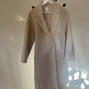 Beige kappa med klassisk krage - Stilren beige kappa med klassisk krage och rak passform. Perfekt för dig som gillar minimalistisk och tidlös stil. Lite nopprig på vissa ställen