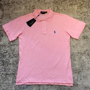 Rosa pikétröja från Polo Ralph Lauren - Snygg rosa pikétröja från Polo Ralph Lauren i bomull med klassisk krage, korta ärmar och blå broderad logga på bröstet. Modellen är slim fit och har knappar framtill. Perfekt för en fräsch och sportig look.