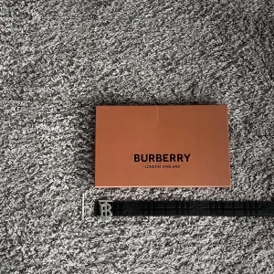 Burberry vändbart bälte  - Säljer ett vändbart Burberry-bälte med ikoniskt TB-spänne i silver. Ena sidan är svart med diskret rutmönster och präglad logga, andra sidan har klassiskt beige rutigt Burberry-mönster med röda och svarta detaljer. Bältet är i skinn och levereras med originalask.