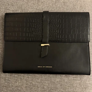 Svart datorfodral från Ideal of Sweden - Stilren svart datorfodral från Ideal of Sweden med croco-präglad ovandel och snyggt spänne i metall. Väskan är tillverkad i skinnimitation och har ett rymligt fack med plats för laptop eller dokument. Perfekt för dig som gillar minimalistisk design med en twist. Men den är lite trasig på sidorna, det syns inte mycket och man behöver inte tänka på de.