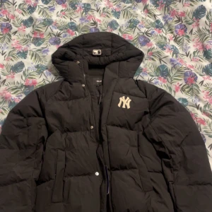 Svart pufferjacka MLB Yankees M - Svart pufferjacka från MLB med New York Yankees broderat på bröstet och ryggen. Jackan har huva, stora fickor och dragkedja med tryckknappar. Perfekt för dig som gillar sportig streetstyle och vill hålla dig varm under kalla dagar. Pris går att diskutera!!