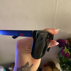 Svarta Gucci solglasögon med röd/grön detalj - Snygga svarta solglasögon från Gucci med klassisk fyrkantig form. Bågarna har en ikonisk röd och grön randig detalj på sidorna och Gucci-logga. Glasen är mörka och bågarna är tillverkade i syntetmaterial. Perfekt accessoar för dig som vill sticka ut.