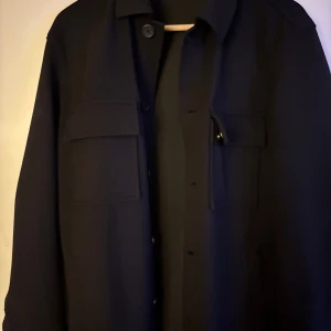 Svart overshirt med fickor från Zara - Stilren svart overshirt med klassisk krage och stora bröstfickor med lock. Jackan har knäppning framtill med knappar och raka ärmar. Perfekt för lager-på-lager och enkel att matcha till olika outfits.