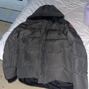 Canada Goose macmillan - Säljer en mörkgrå jacka från Canada Goose med huva och klassisk logga på ärmen. Jackan har quiltad design, dragkedja och stora fickor framtill. Perfekt för kalla dagar och har en clean look som är riktigt nice.