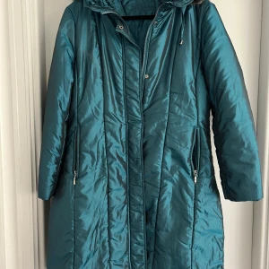 Grön/blå  pufferjacka med huva - Säljer en blå/grön  pufferjacka från Oneness med lång passform och huva med fuskpäls. Jackan har dragkedja och tryckknappar framtill, samt två fickor med dragkedja och en innerficka. Praktisk, lättburen, snygg quiltad look.