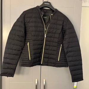 Svart dunjacka från Rockandblue - Svart dunjacka från Rockandblue med guldfärgade dragkedjor fram, vid fickor och ärmslut. Jackan har quiltad design och är tillverkad i polyester med äkta dunfyllning. Snygg, stilren och passar perfekt till kyliga dagar.