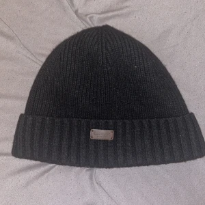 Svart ribbad mössa från Barbour - Svart ribbstickad mössa från Barbour med uppvikt kant och en liten metallbricka framtill med logotyp. Klassisk och stilren modell som passar perfekt till kyliga dagar.