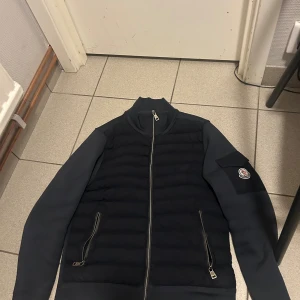 Svart pufferjacka från Moncler S(BUD) - Svart pufferjacka från Moncler i storlek S. Jackan har quiltad front, släta ärmar och Moncler-logga på vänster ärm. Dragkedja framtill och två sidofickor med dragkedja. Hög krage och stilren design, perfekt för dig som gillar exklusiva streetwear-vibbar.