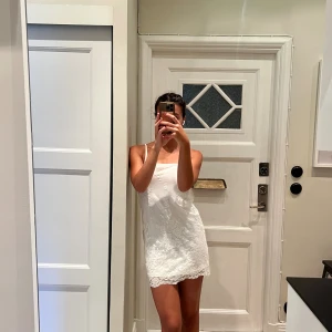 Vit spetsklänning från Ida Sjöstedt - Söt vit kort klänning från Ida Sjöstedt med tunna axelband och blommig spets.. Sidenliknande material under spetsen och fina spetsdetaljer längs nederkanten. Perfekt som studentklänning! 