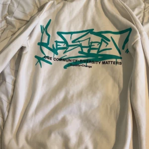 Vit hoodie med grön graffiti-tryck - Vit hoodie med grön graffiti-inspirerad text och budskapet 'One Community - Diversity Matters' på ryggen. Har huva med snörning och känguruficka framtill. Skön och avslappnad passform Säljer båda tsm för 750 både tröjan o jeans är helt ny men utan prislap men det går att köpa tröjan själv och byxan