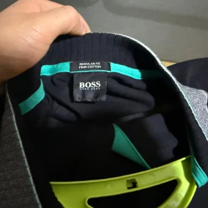 Svart pikétröja från Hugo Boss - Svart pikétröja från Hugo Boss i mjuk pima-bomull. Tröjan har turkosa detaljer vid knäppningen och logga på bröstet samt gråmelerad krage med turkos kant. Klassisk piké med korta ärmar och stilren design.