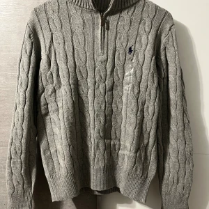 Polo Ralph Lauren half zip - Orginal pris: 2113kr, men endast 899kr hos Luxe Vibes! Kabelstickad grå tröja från Polo Ralph Lauren med halv dragkedja och broderad marinblå logga på bröstet. Tröjan har ribbade muddar och hög krage, perfekt för lager-på-lager. Klassisk och stilren design i mjukt stickat material. 