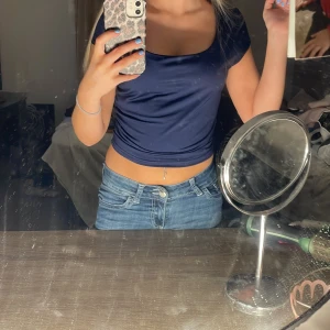 Mörkblå croppad topp med kort ärm - Säljer en stilren mörkblå croppad topp med korta ärmar och rundad fyrkantig halsringning. Toppen har en tight passform och är tillverkad i ett mjukt, glansigt syntetmaterial som sitter snyggt mot kroppen. Perfekt för dig som gillar enkla men trendiga plagg.