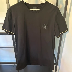 Svart t-shirt från Armani Exchange - Svart t-shirt från Armani Exchange med liten logopatch på bröstet. Klassisk rund hals och korta ärmar med gul detalj på ärmsluten. Tillverkad i mjuk bomull, perfekt för en clean och stilren look.