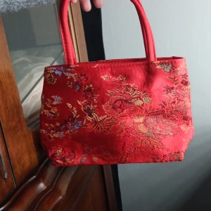 Röd handväska med blommigt mönster - Snygg röd handväska med blommigt mönster i guld och blått. Väskan har två handtag och är tillverkad i sidenliknande material. Insidan är ljus och har dragkedja upptill. Perfekt för att addera färg och stil till din outfit.