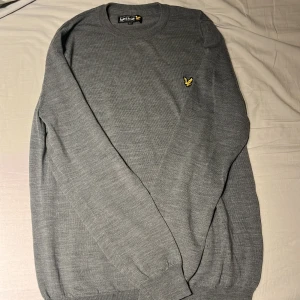 Grå stickad tröja från Lyle & Scott - Säljer en stilren grå stickad tröja från Lyle & Scott med rund halsringning och klassisk gul logga på bröstet. Tröjan har lång ärm och är perfekt för lager-på-lager under kyliga dagar. Den är i storlek L men passar även M. Priset går att diskutera