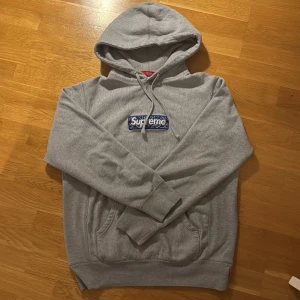 Grå Supreme hoodie med blå box logo - Supreme hoodie i grått med klassisk blå box logo på bröstet. Hoodien har snörning i huvan, känguruficka och ribbade muddar. Box logon är broderad med vitt Supreme-tryck och paisleymönster. Tillverkad i mjuk bomull för maximal komfort.