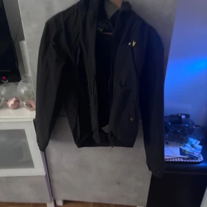 Svart vindjacka från Lyle & Scott - Svart vindjacka från Lyle & Scott med huva och dragkedja framtill. Jackan har en klassisk passform och är tillverkad i polyester. Liten gul logga på bröstet ger en clean look. Perfekt för blåsiga dagar.