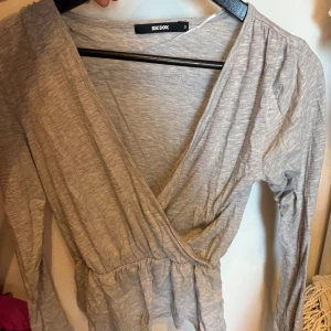 Grå omlottopp från BikBok - Säljer en ljusgrå omlottopp från BikBok i mjuk bomullsmix. Toppen har lång ärm, v-ringning och smockad midja som ger en snygg siluett. Perfekt att styla med jeans eller kjol för en chill look.