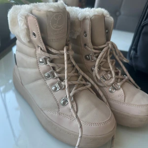 Beige vinterkängor med fuskpäls - Säljer ett par beige vinterkängor från Linears med fluffig fuskpäls runt skaftet och snörning med metallhakar. Skorna har platt sula och är tillverkade i syntetmaterial med mockakänsla. Perfekta för kalla dagar och har en snygg, chunky look.